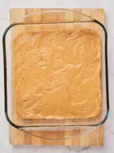 A square glass baking dish filled with a smooth, light peanut butter batter, placed on a wooden cutting board.