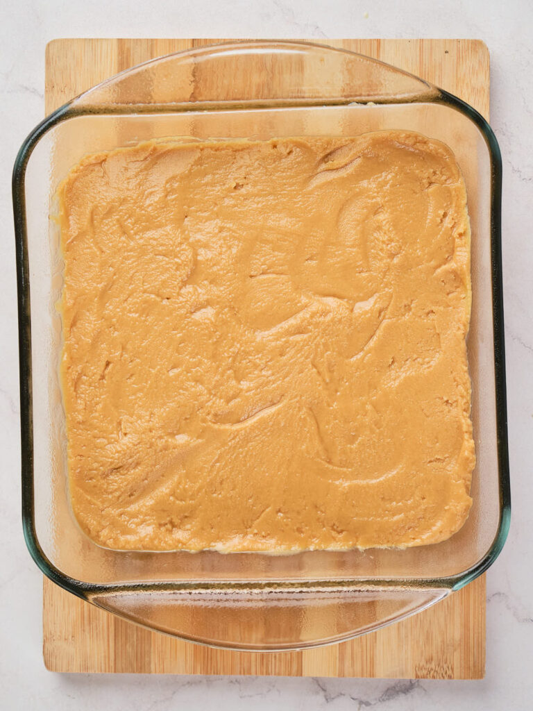 A square glass baking dish filled with a smooth, light peanut butter batter, placed on a wooden cutting board.