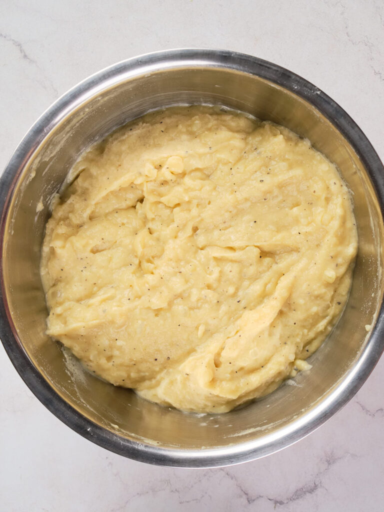 A bowl of mashed potatoes in a stainless steel bowl on a light-colored surface.