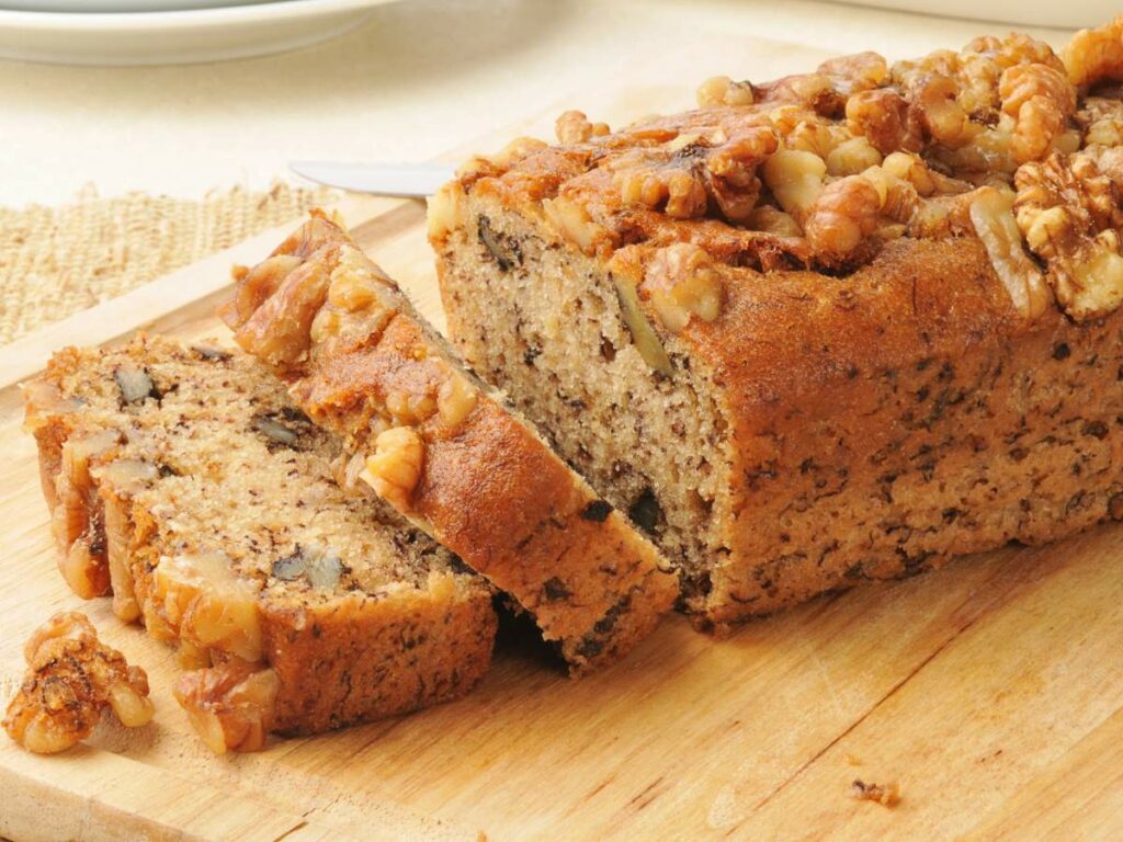 A loaf of bread sliced on a wooden cutting board, showing a moist interior with chunks of nuts throughout.