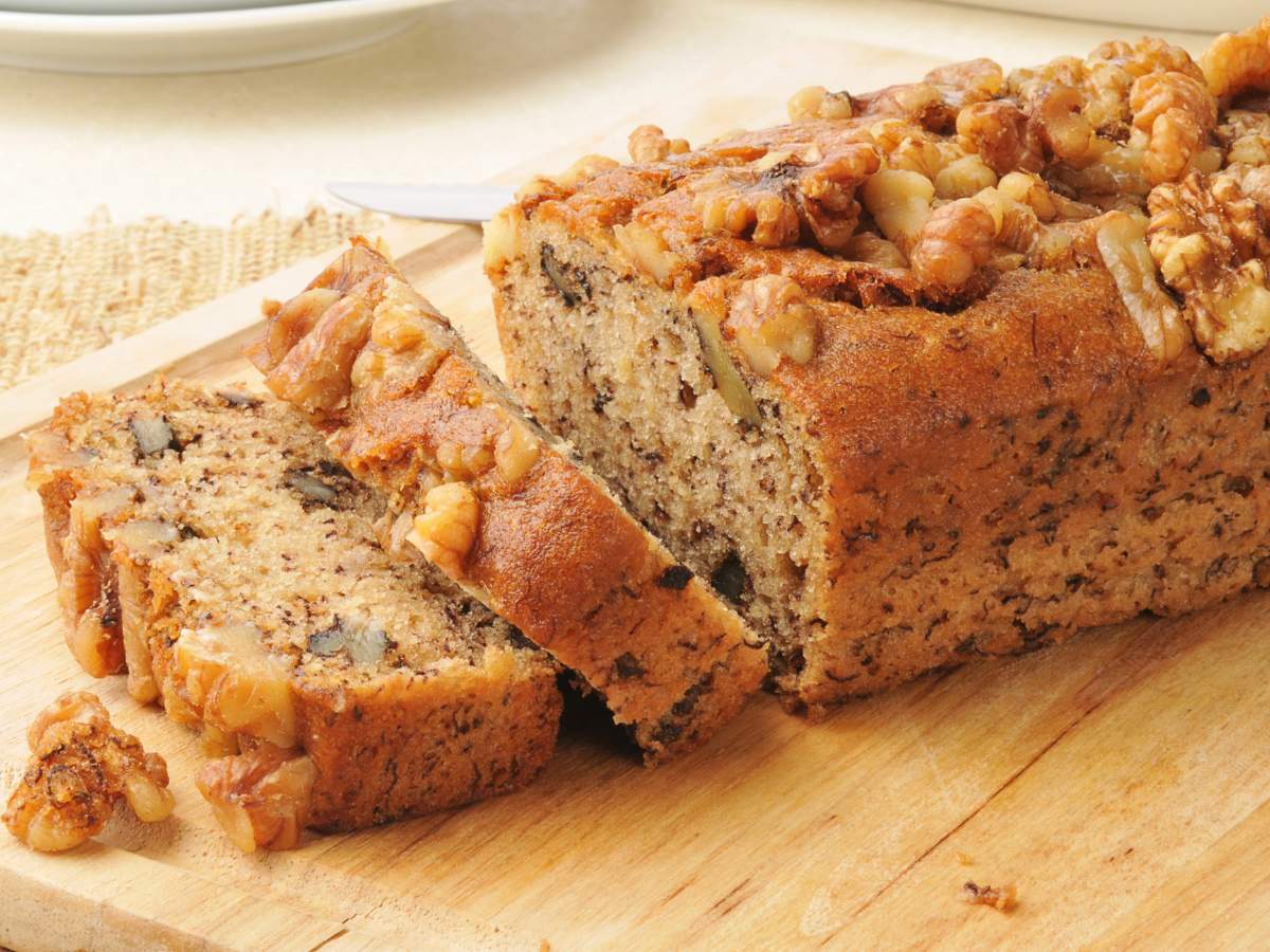 A loaf of bread sliced on a wooden cutting board, showing a moist interior with chunks of nuts throughout.