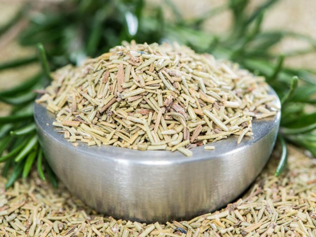 A metal bowl filled with dried rosemary sits atop a surface.