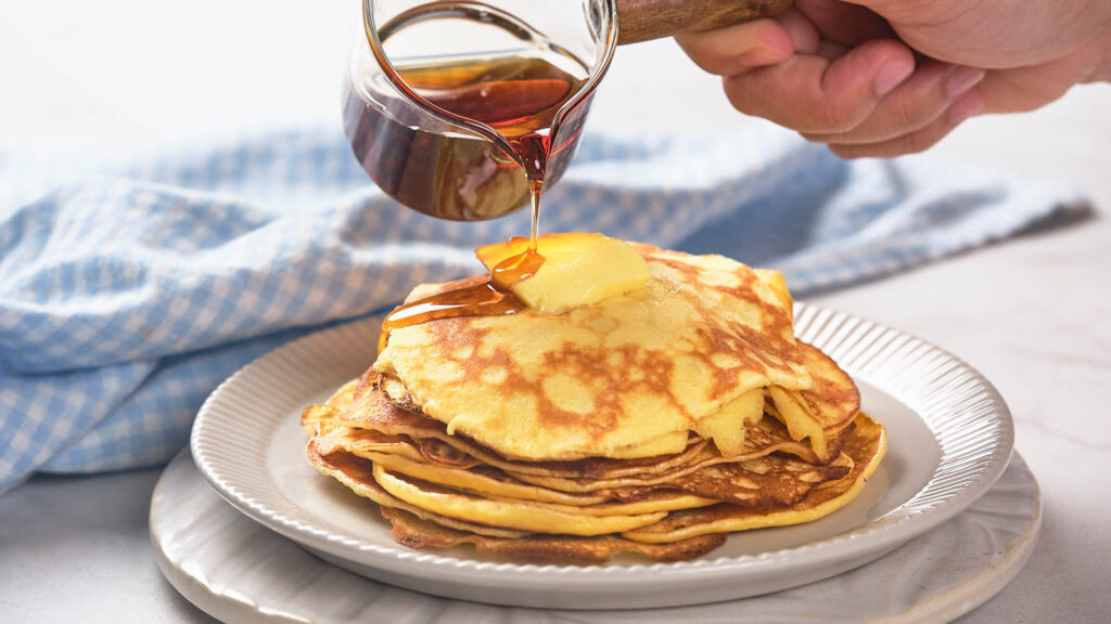 A stack of pancakes topped with butter is being drizzled with syrup.