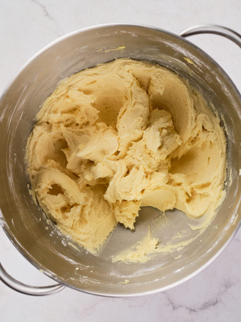 A metal mixing bowl containing creamy, yellow cookie dough on a light marble surface.