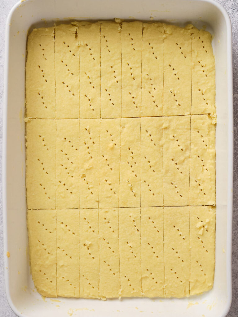 A rectangular baking dish filled with unbaked shortbread dough, cut into squares with fork holes.