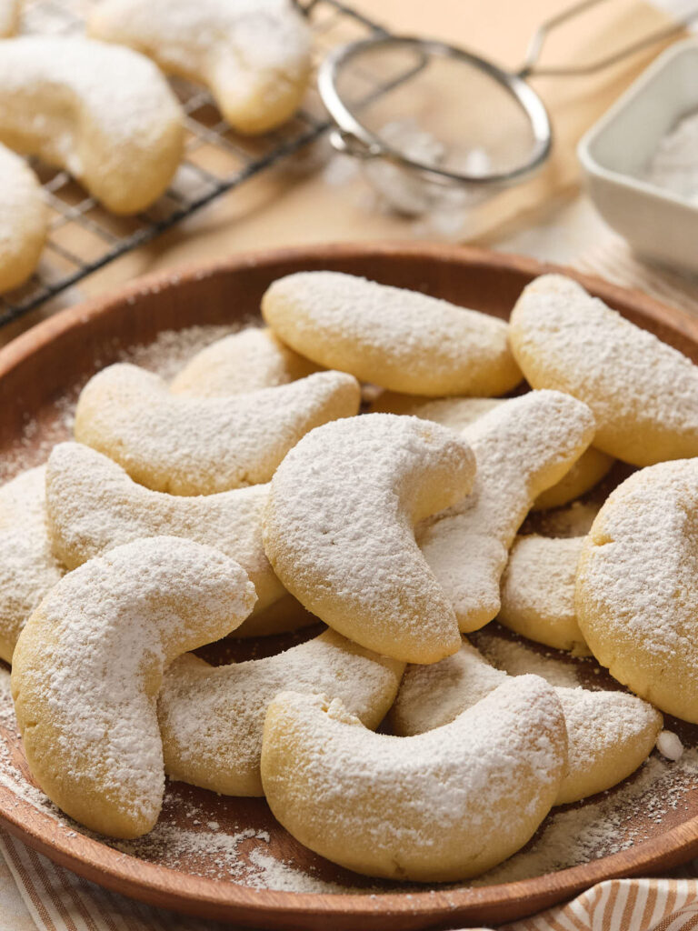 A wooden plate filled with almond crescent cookies dusted with powdered sugar.