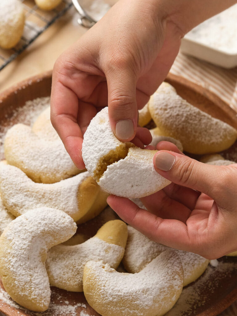 Hands holding and breaking a crescent-shaped cookie dusted with powdered sugar.