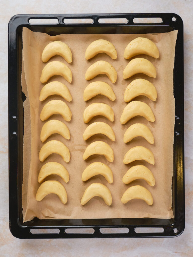 A baking tray lined with parchment paper holds twenty unbaked, crescent-shaped cookies arranged in four rows.