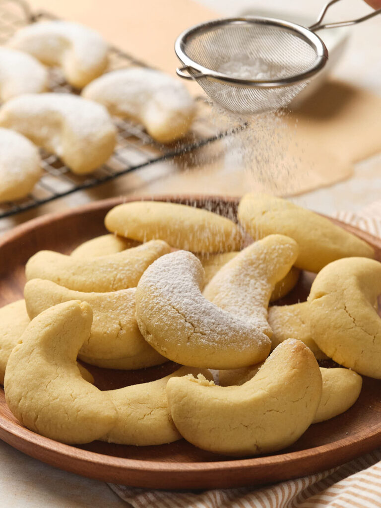 A plate of almond crescent cookies is being dusted with powdered sugar.