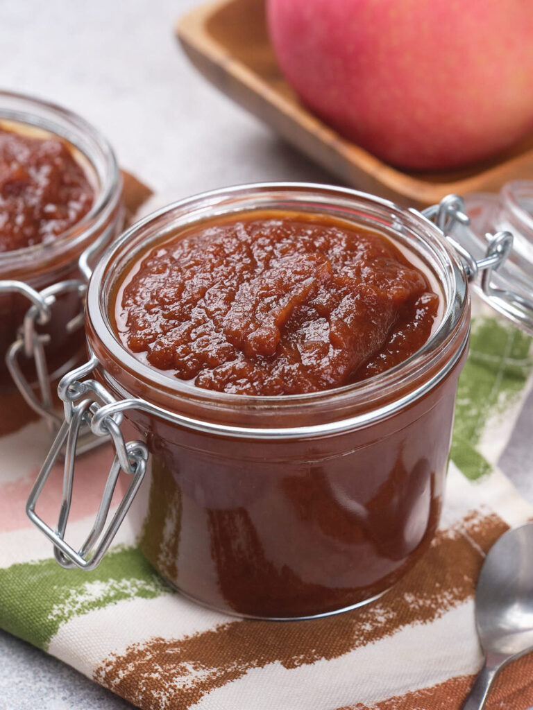 A jar of apple butter on a patterned cloth, with a spoon and an apple in the background.