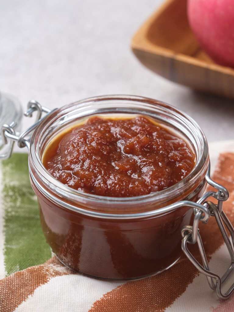 A glass jar filled with apple butter sits on a striped cloth, with an apple in the background.