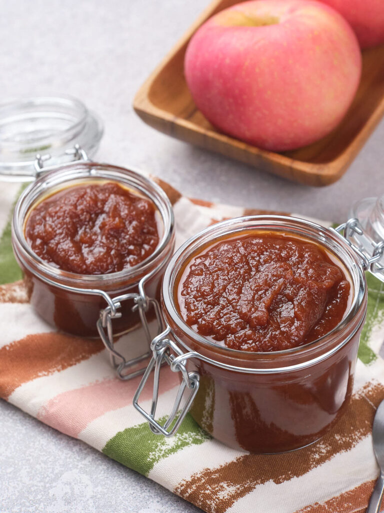 Two glass jars filled with apple butter sit on a striped cloth, with fresh apples in a wooden tray nearby.