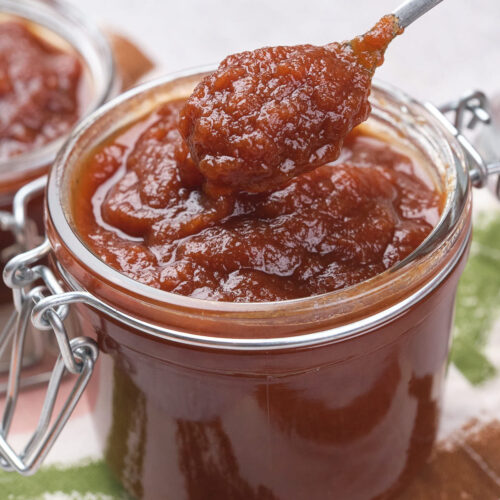 A jar of thick, brown apple butter with a spoon scooping some out.