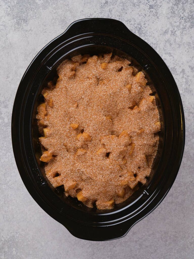 Slow cooker with cinnamon-sugar coated apple dessert on a gray surface.