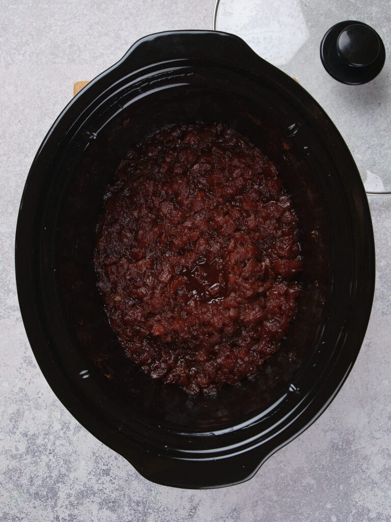 Slow cooker with cooked apple butter inside, placed on a light gray surface.