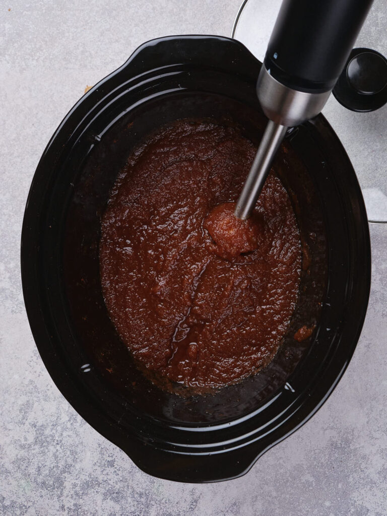 Immersion blender mixing thick brown sauce in a slow cooker on a gray countertop.