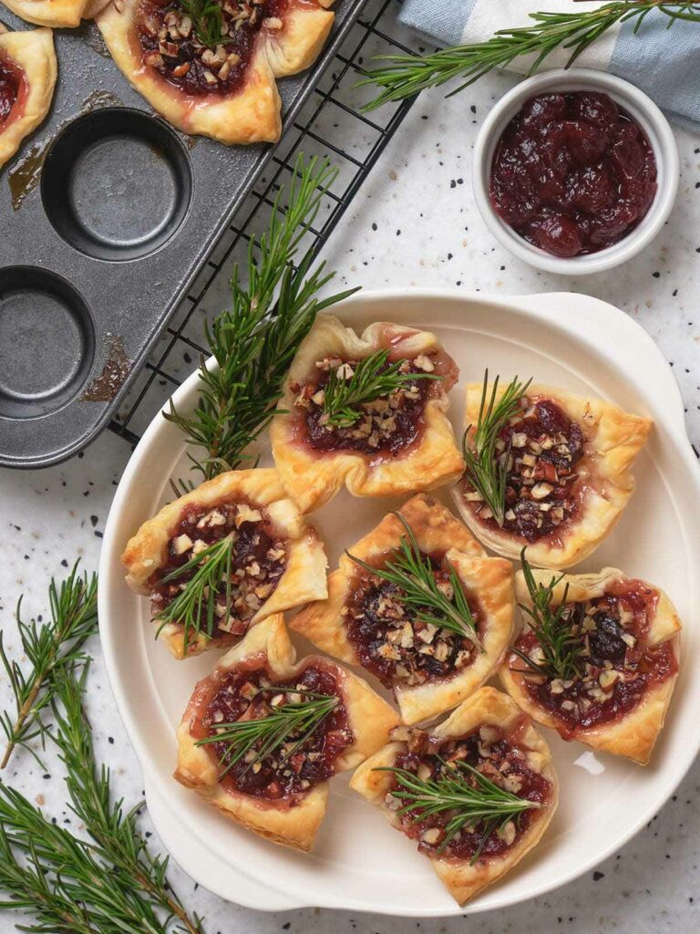 Brie bites topped with cranberry sauce, nuts, and rosemary on a white plate.