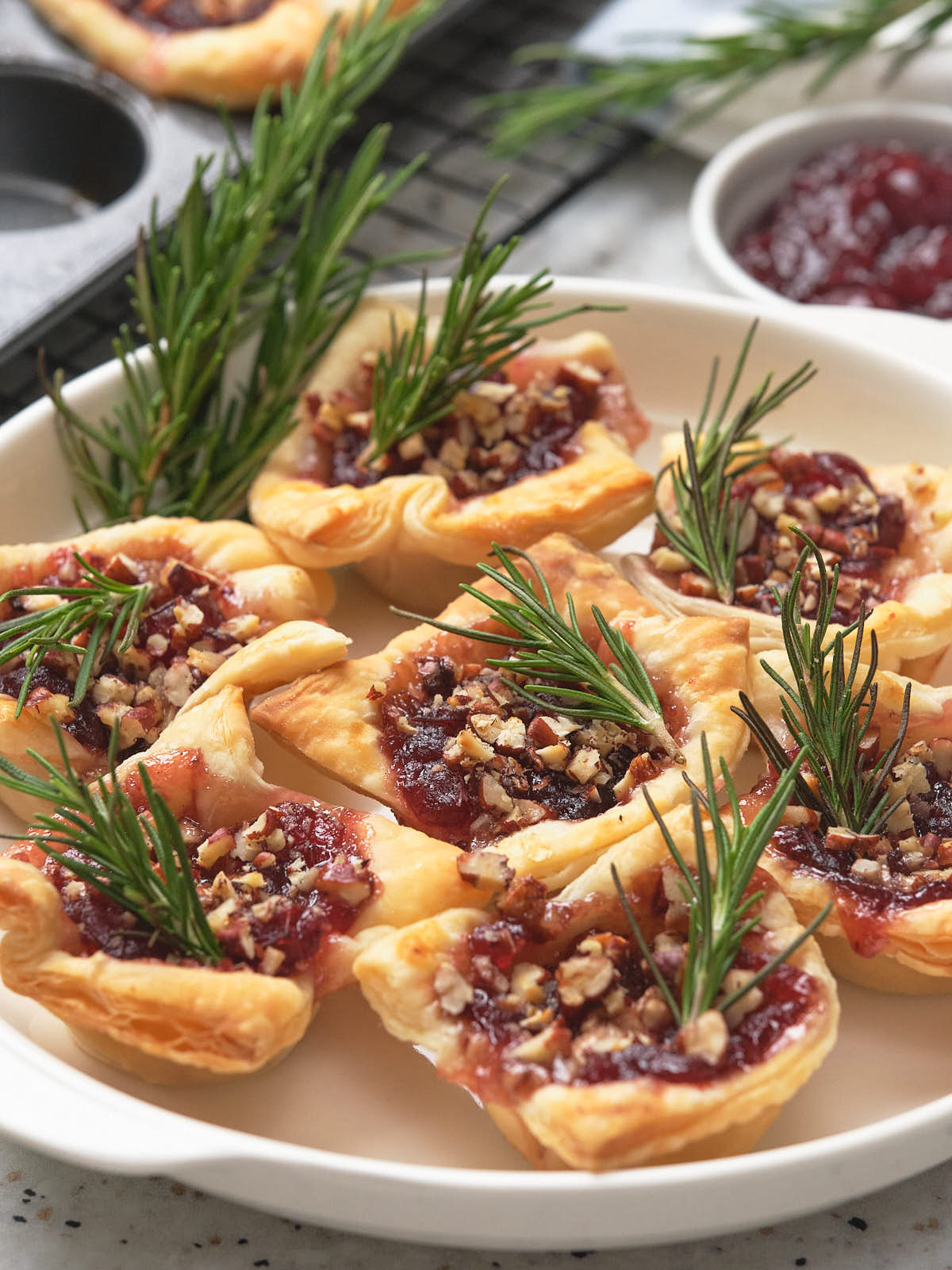 Brie bites topped with cranberry sauce, chopped nuts, and garnished with rosemary on a white plate.