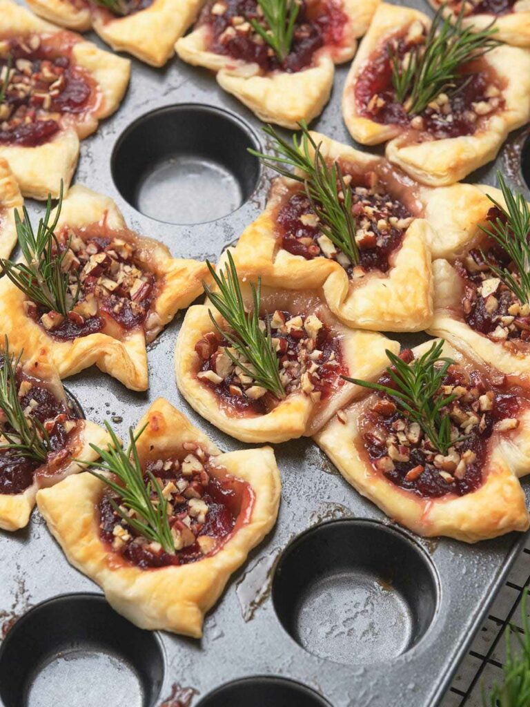 Brie bites with cranberry sauce, chopped nuts, and rosemary sprigs in a muffin tray.