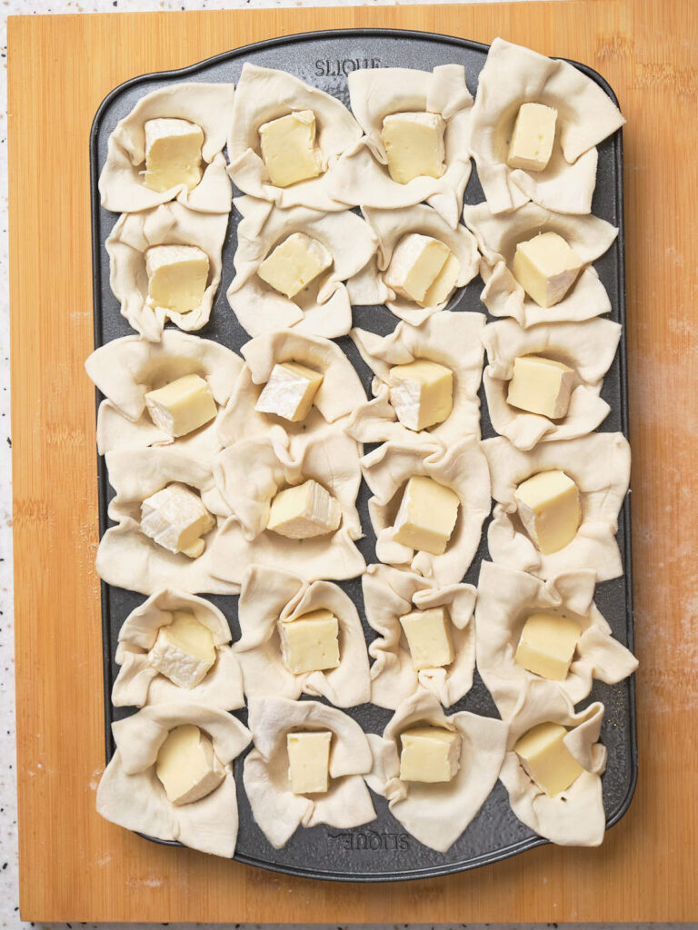 A baking tray with unbaked pastry squares, each filled with a cube of cheese.