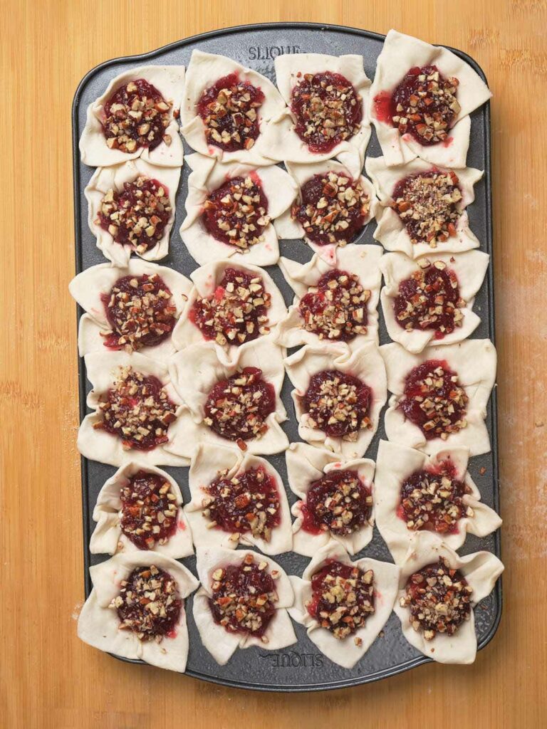 A baking tray filled with unbaked pastries, each topped with a red fruit filling and chopped nuts.