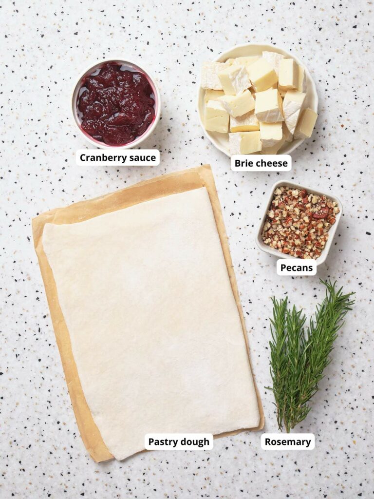 Pastry dough, brie cheese, cranberry sauce, pecans, and rosemary arranged on a speckled surface.