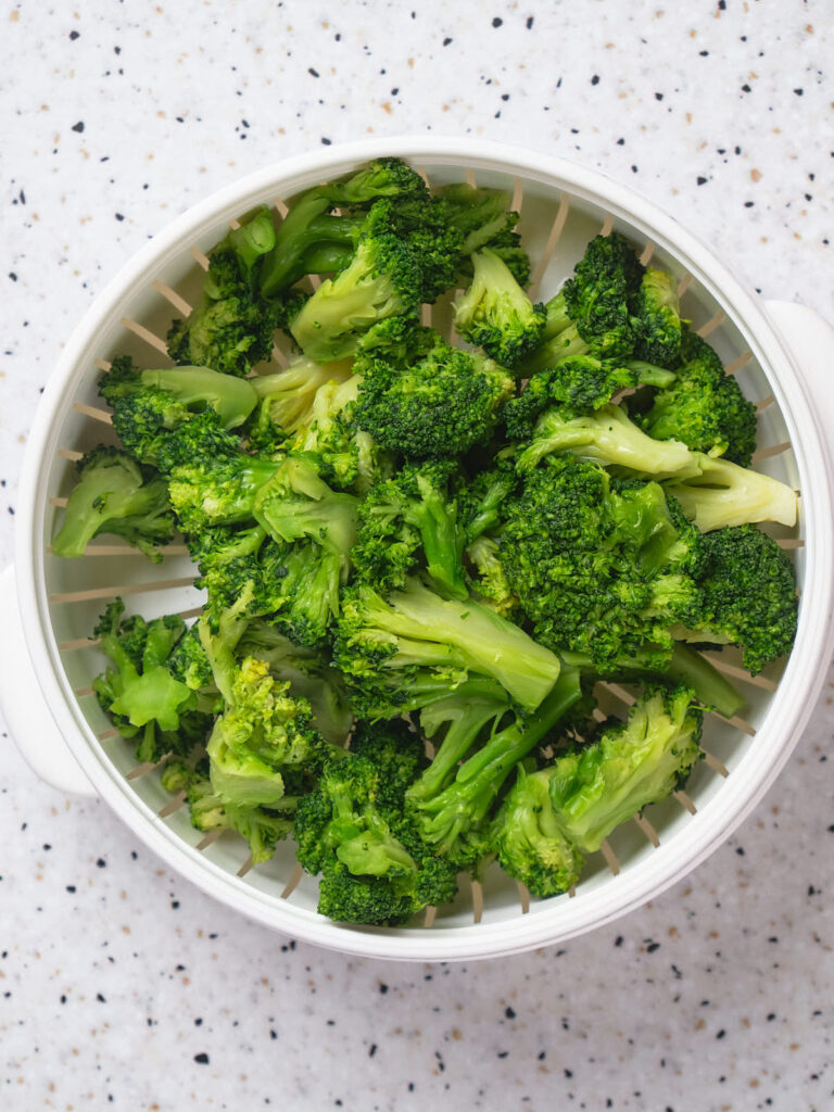 Steamed broccoli florets in a white colander on a speckled countertop.