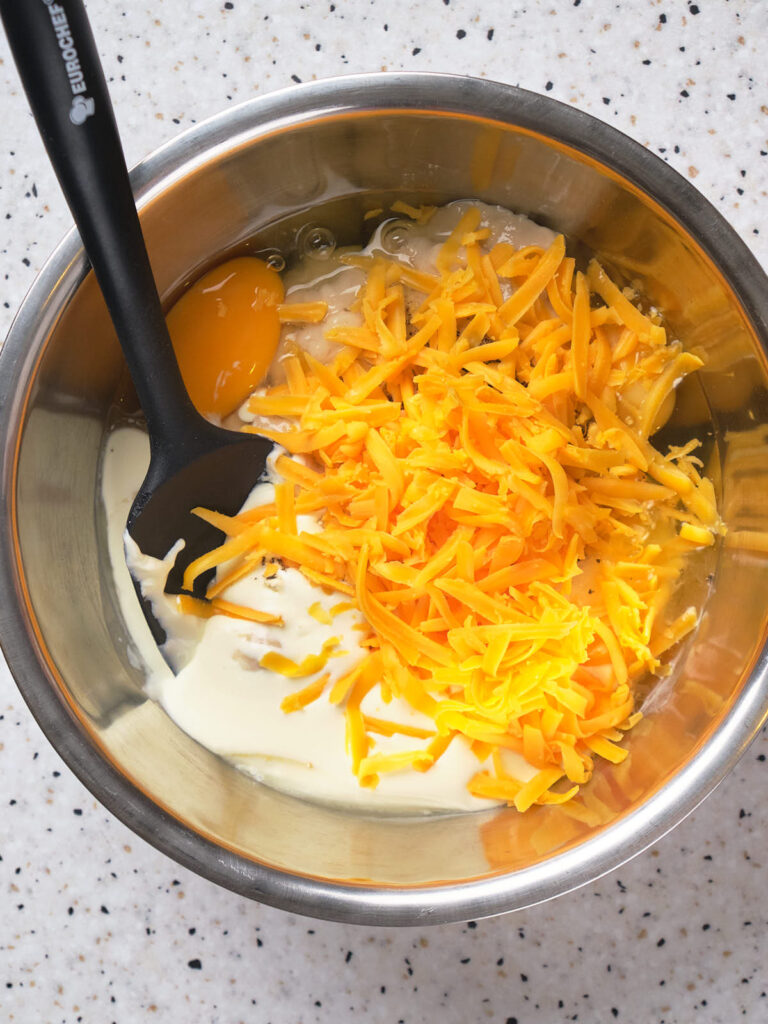 A mixing bowl with shredded cheddar cheese, eggs, and cream being stirred with a black utensil.