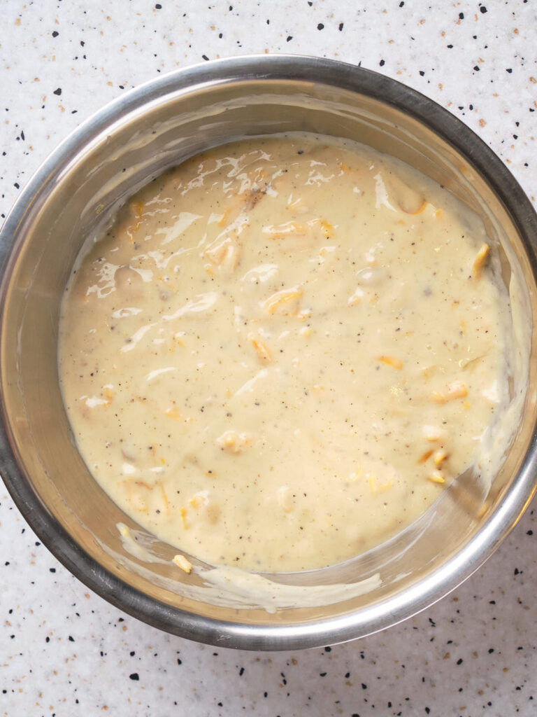 A stainless steel bowl containing a creamy batter mixture with visible shreds of cheese, placed on a speckled countertop.