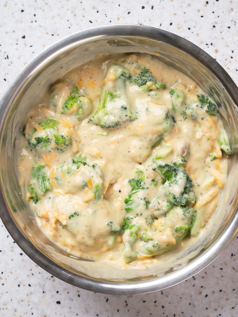 A metal bowl filled with broccoli and shredded cheese mixed into a creamy batter.