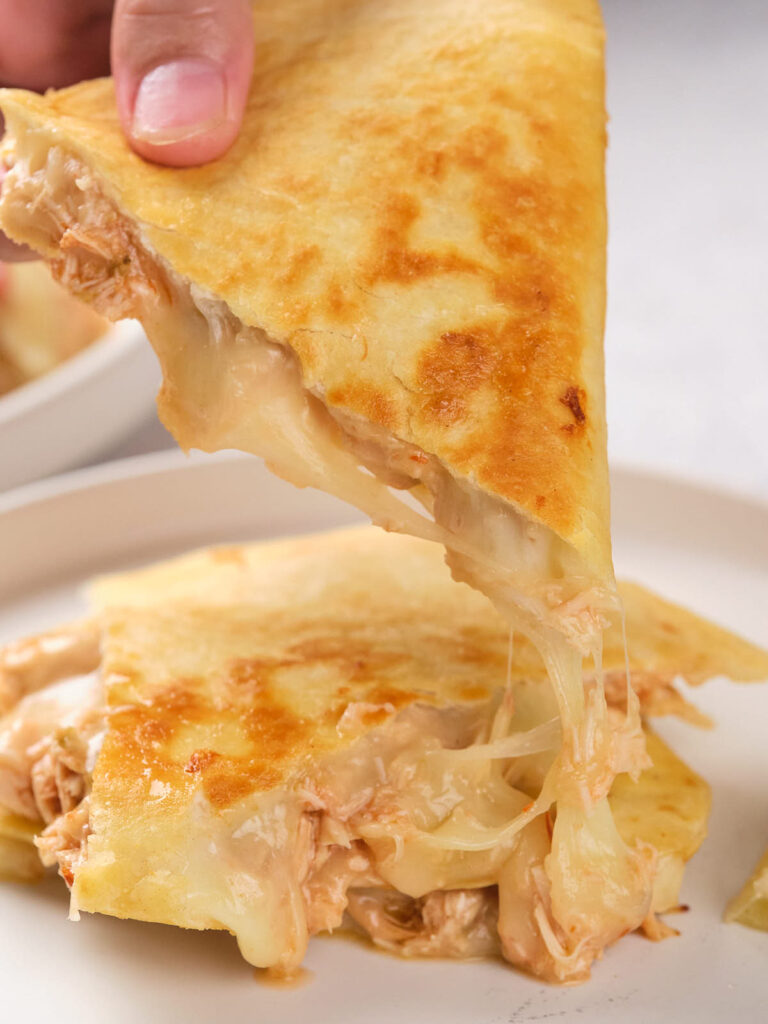 A hand lifts a slice of a cheesy chicken quesadilla from a plate.