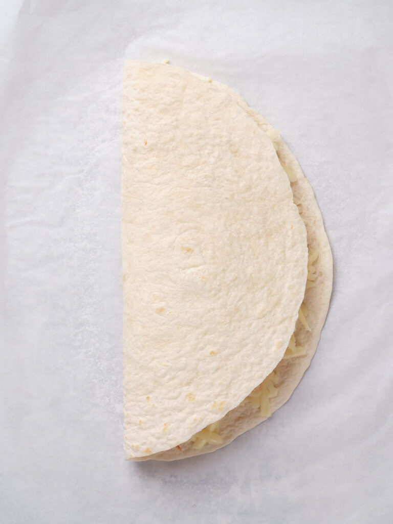 A folded flour tortilla on white parchment paper, partially revealing cheese filling.