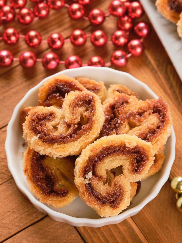 A bowl of cinnamon palmiers with a swirled filling on a wooden table, surrounded by red beads.