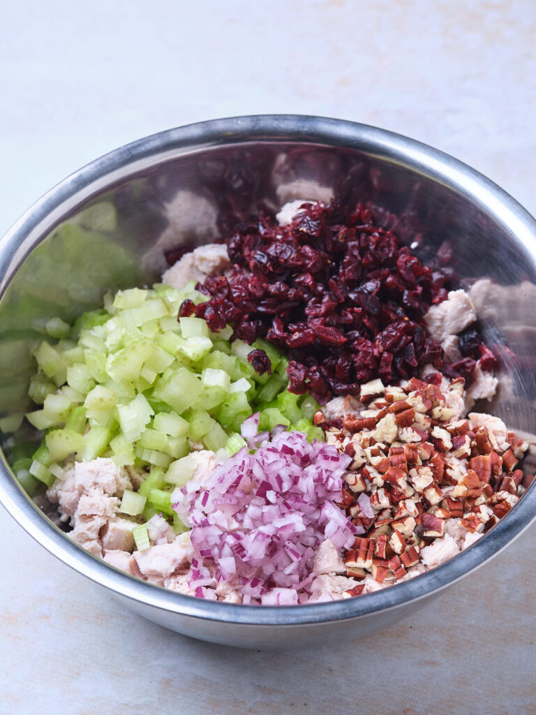 A metal bowl contains diced celery, chopped cranberries, minced red onion, and chopped nuts on top of chunks of cooked chicken.