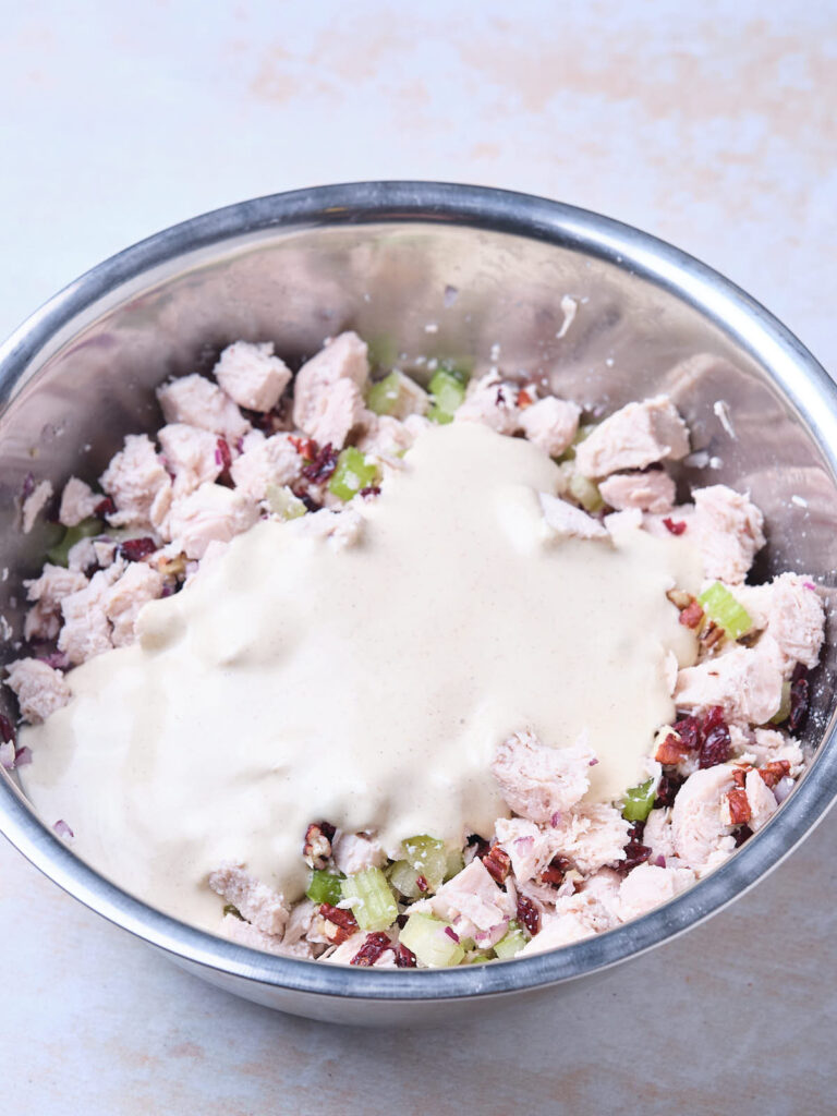 A stainless steel bowl containing a mixture of chopped chicken, celery, cranberries, and pecans. A light cream-based dressing is poured over the mixture.