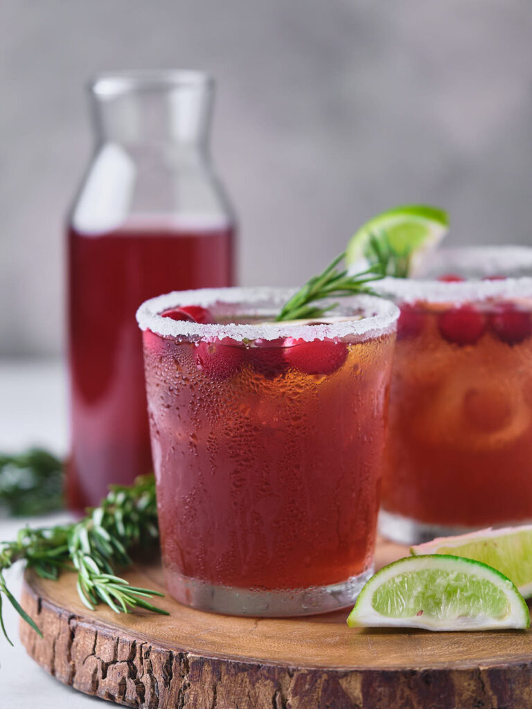 Two glasses of a cold cranberry margarita and lime slices sit on a wooden board.