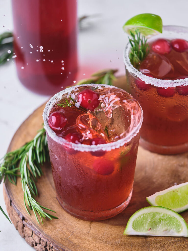 Two glasses of cranberry margarita with ice, garnished with cranberries and lime wedges.