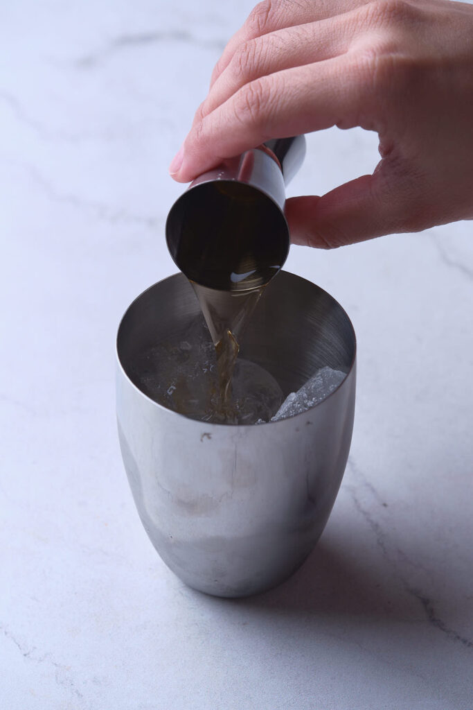 A person pours a brown liquid from a jigger into a metal shaker filled with ice on a marble surface.