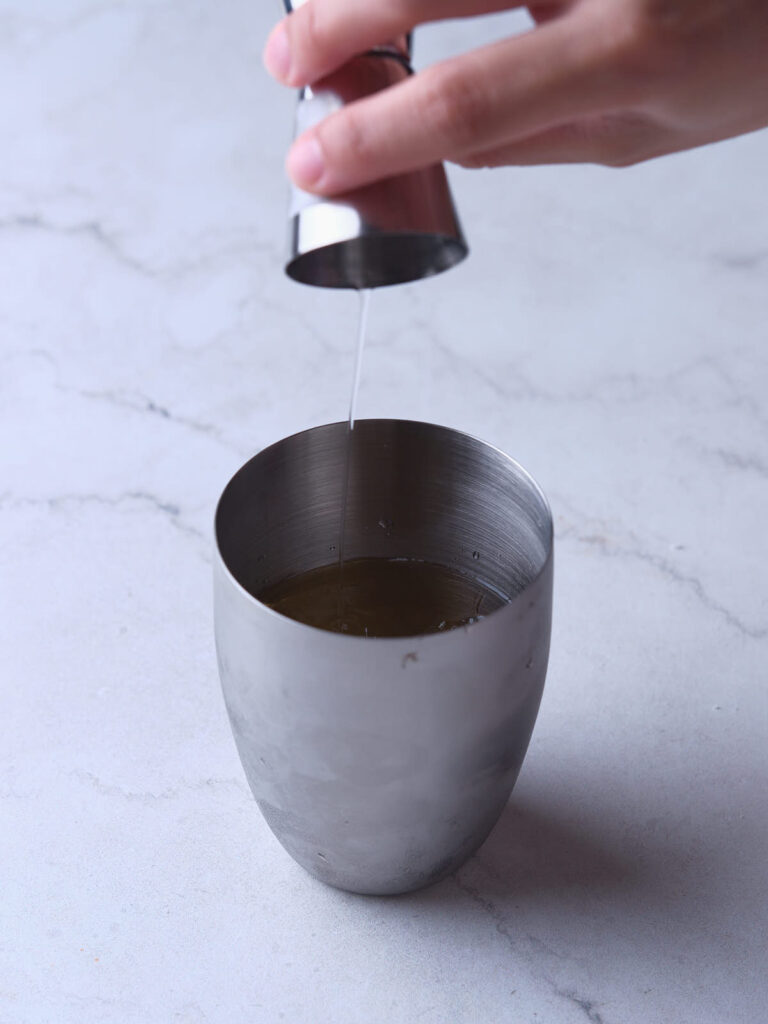 A hand pours liquid from a jigger into a stainless steel cup on a marble surface.