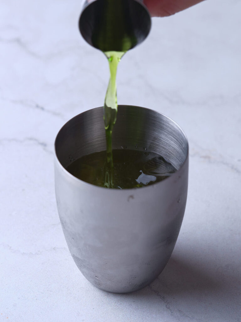 A hand pours green liquid into a gray steel cup on a white marble surface.