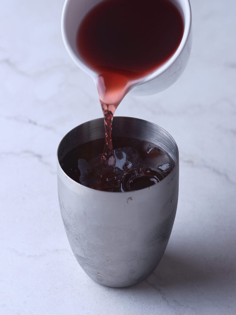 A hand pours a red liquid from a small white cup into a silver cup filled with ice on a light gray surface.