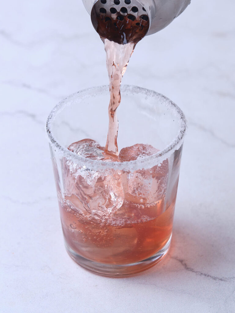 A pink-colored cocktail being poured from a shaker into a glass with ice cubes and a sugar-rimmed edge.