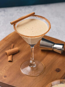 Eggnog martini in a glass with a cinnamon stick, set on a wooden board with cinnamon sticks nearby.