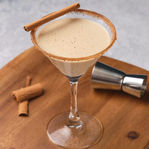 Eggnog martini in a glass with a cinnamon stick, set on a wooden board with cinnamon sticks nearby.