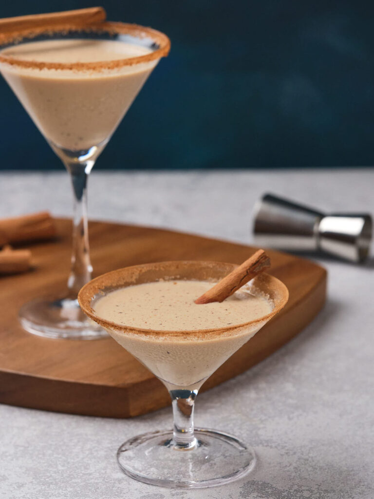 Two glasses filled eggnog martini, garnished with cinnamon sticks and rimmed with cinnamon sugar.