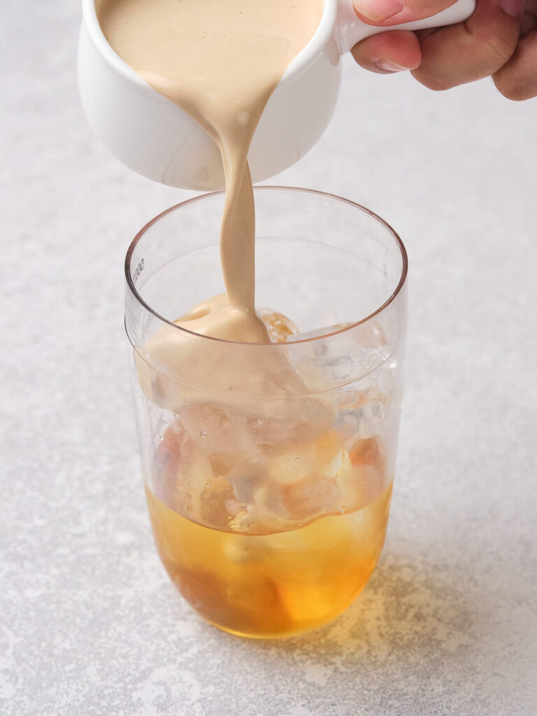 Pouring creamy milk into a glass of iced tea on a light-colored surface.