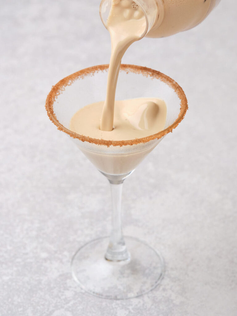 Creamy beverage being poured into a martini glass with a sugared rim on a light gray surface.