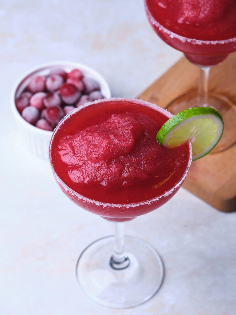 A glass of frozen red cocktail garnished with a lime slice, with a small bowl of frozen cranberries nearby.