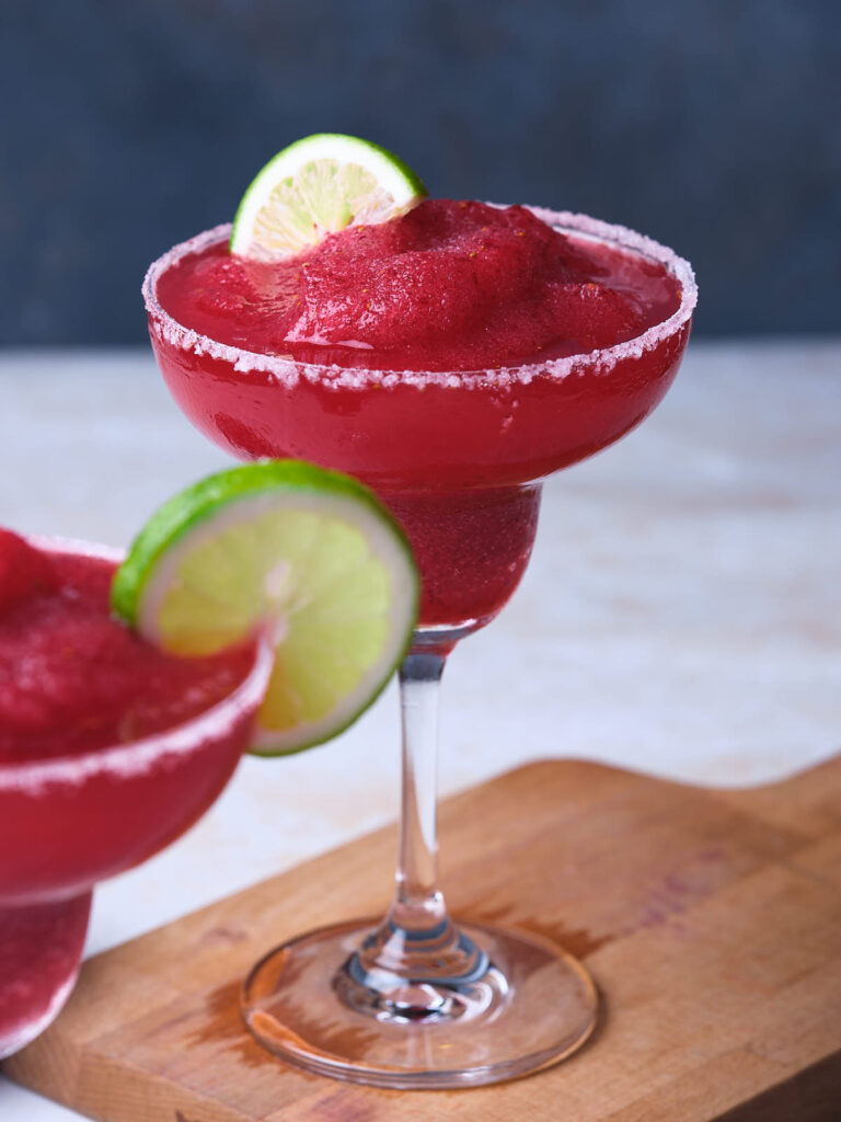 Two glasses of frozen cranberry margarita garnished with lime wedges.