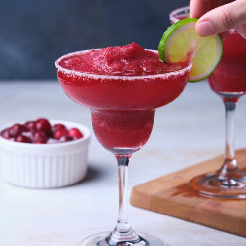 A hand garnishes a frozen cranberry margarita glass with a slice of lime.